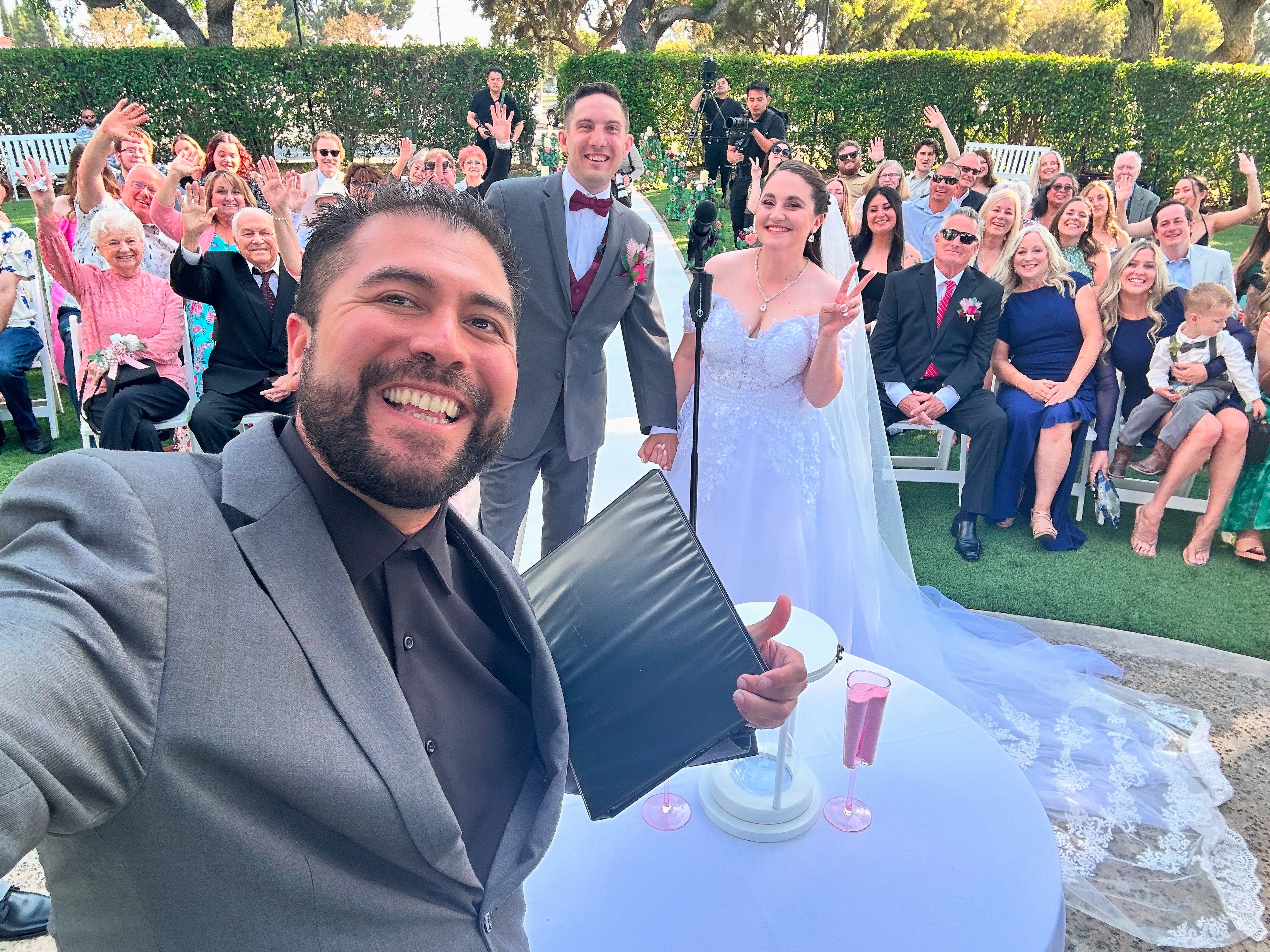 Mike taking a selfie with couple and full crowd waving at an outdoor ceremony