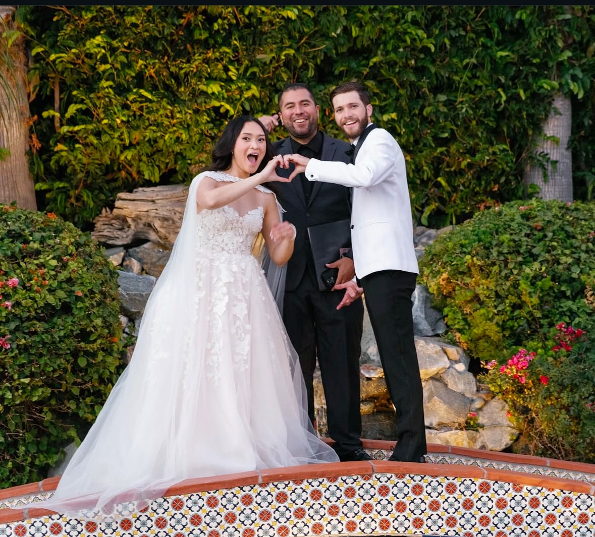 Mike with bride and groom making a heart with their hands in a lush garden