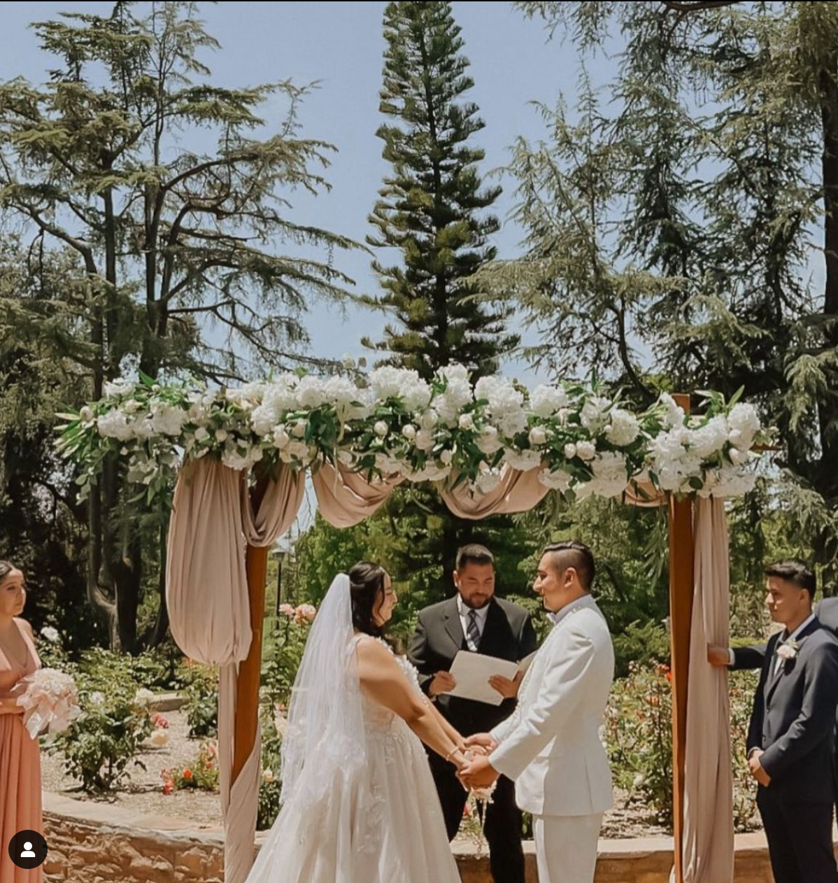 Bride and groom holding hands under a white floral arch in a forest garden